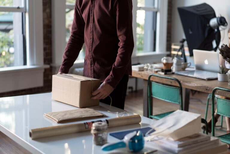 a person in a home office from the shoulders down, taping a cardboard box that sits on a table with other shipping supplies.